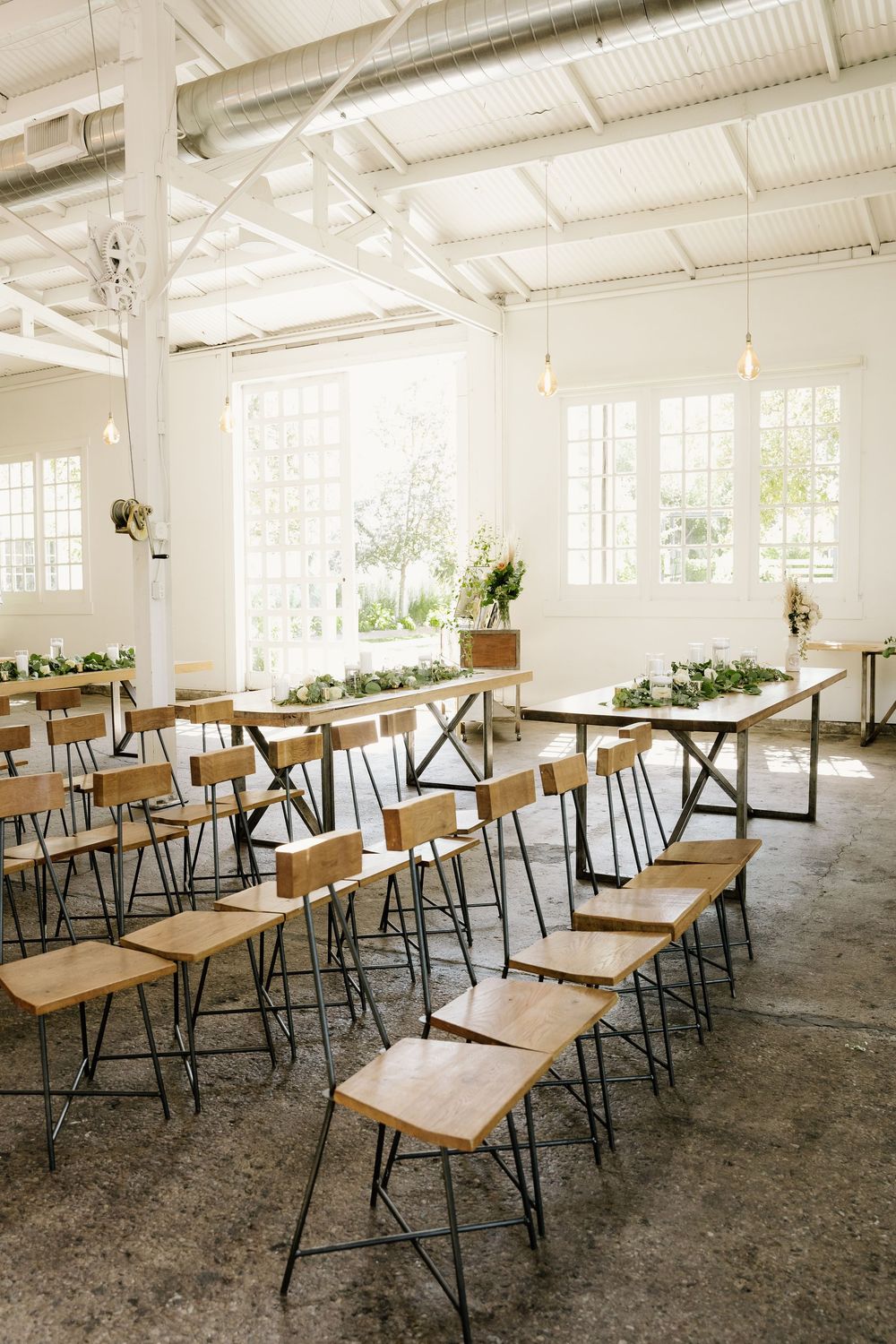 Rows of wooden chairs with metal frames arranged in a bright white industrial venue with large windows and greenery decorations.