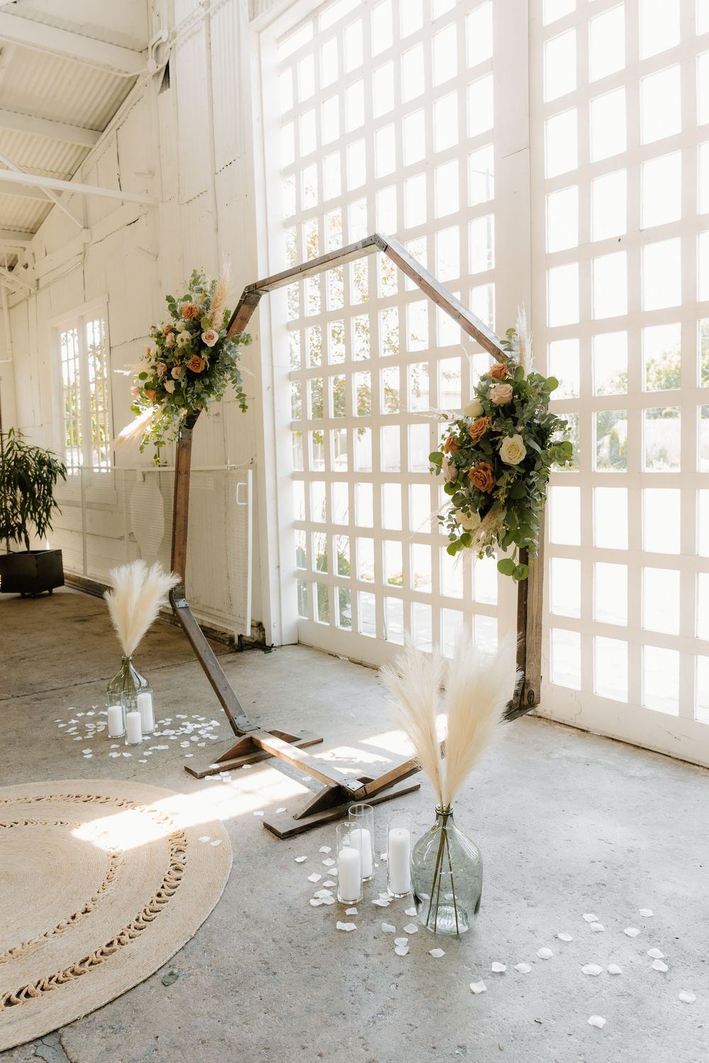 Minimalist wedding ceremony arch with floral arrangements and glass vases against white latticed windows in an industrial space.
