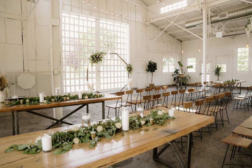 Wedding reception setup with long wooden tables decorated with greenery garlands in white industrial space.