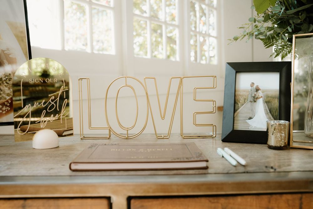 Gold LOVE sign and wedding decor displayed on wooden table near window with florals.
