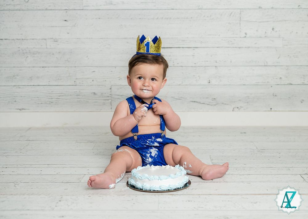 Small child in blue outfit and crown celebrates first birthday with cake on white wooden background.