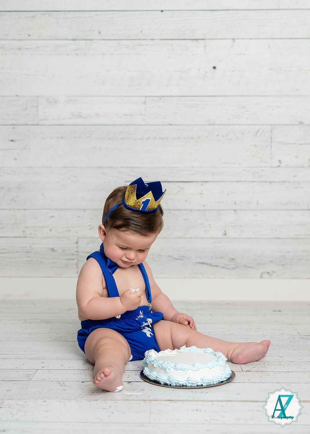 A young child wearing a royal blue outfit and crown sits on a white wooden floor with a birthday cake.