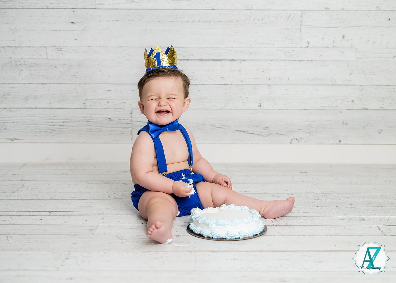 A baby wearing a blue outfit and small crown sits on a white floor with a cake for their first birthday celebration.