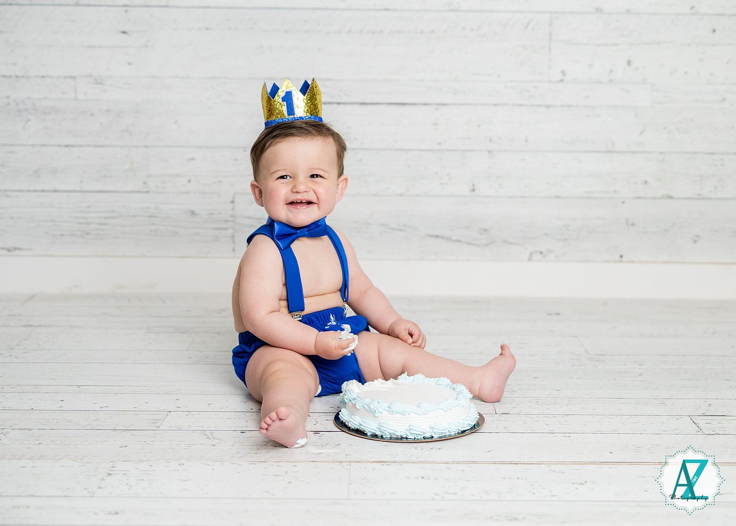 A baby wearing a blue outfit and small crown sits on a white floor with a cake for their first birthday celebration.