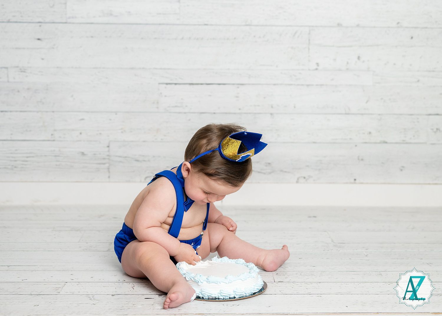Small figure in a blue outfit and bow sits on white wood floor examining a white fluffy cushion prop.