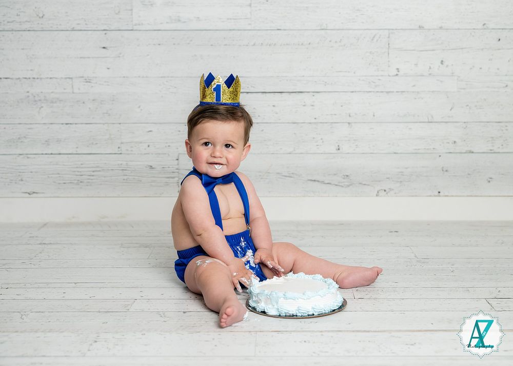 Baby wearing a blue crown and bowtie sits next to a smash cake against a white wooden backdrop for first birthday photos.