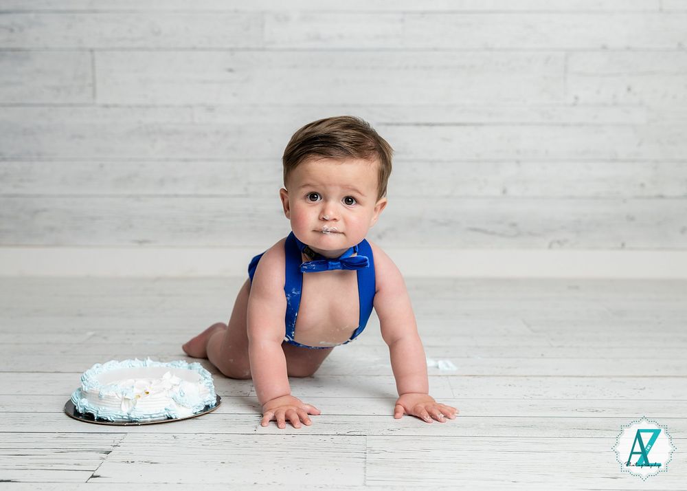 A baby in a blue bowtie and suspenders crawls on a light wooden floor next to a smash cake for their first birthday.