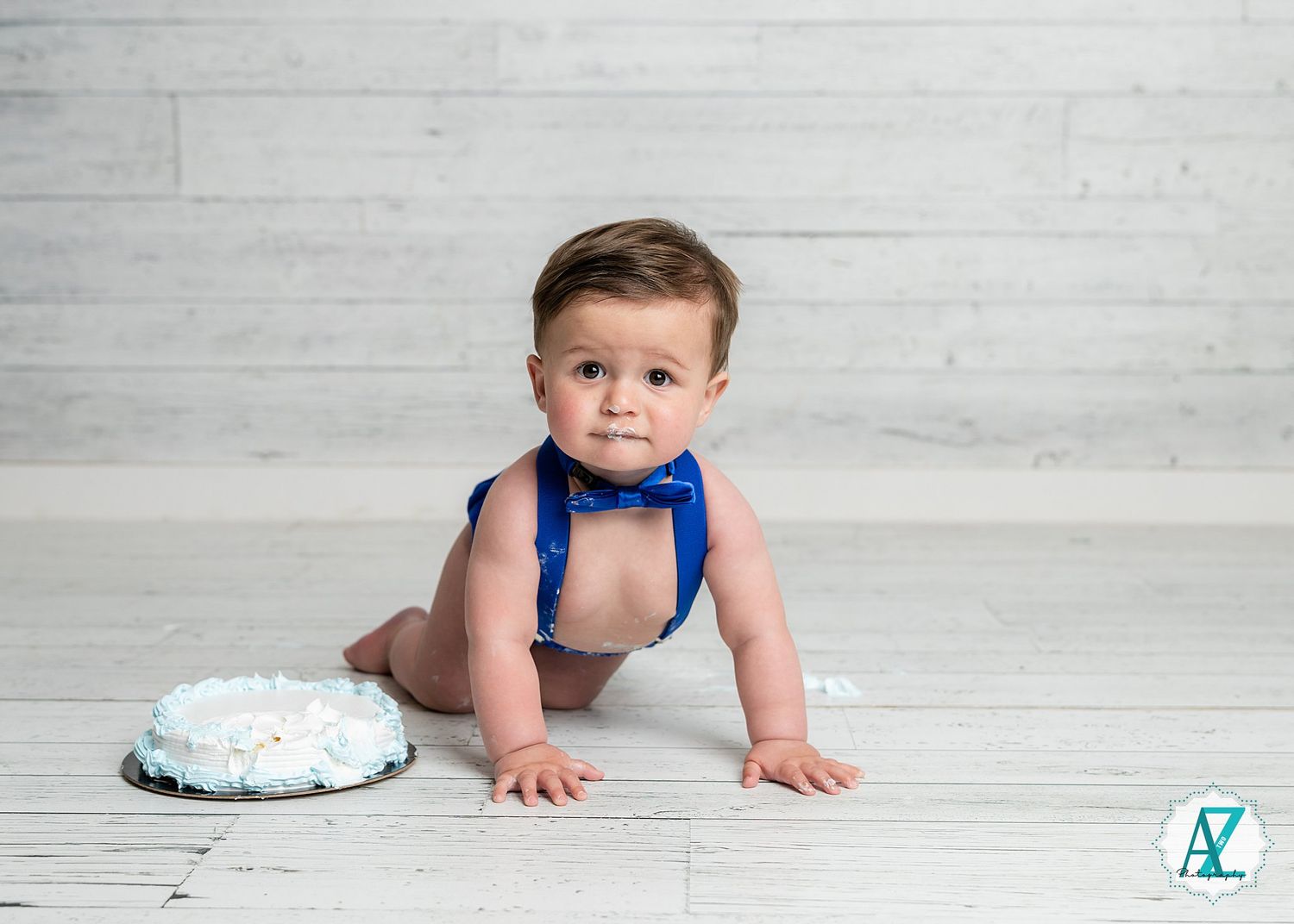 A baby in a blue bowtie and suspenders crawls on a light wooden floor next to a smash cake for their first birthday.