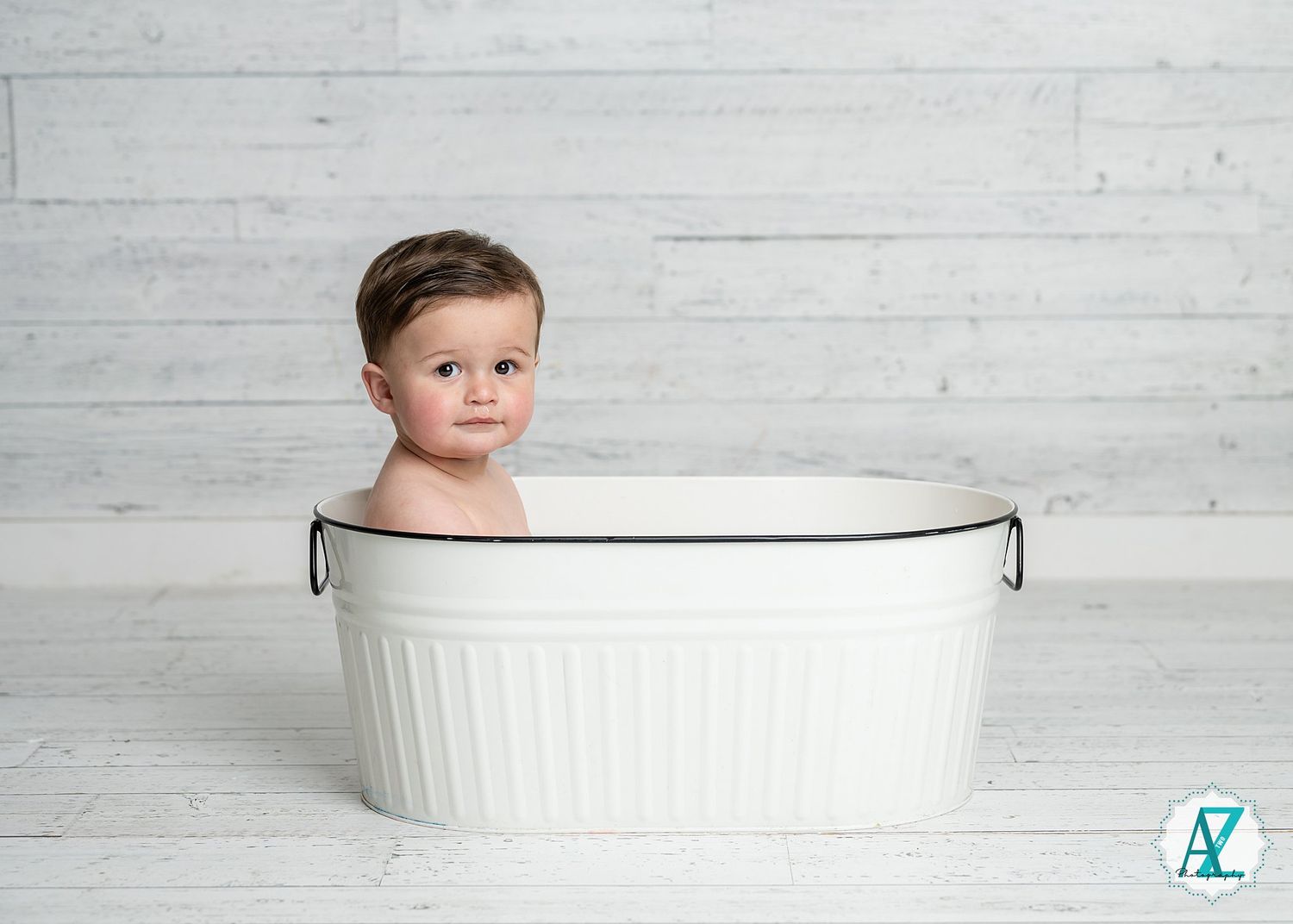 Baby sitting in a white vintage-style oval wash basin against a light wooden plank backdrop.