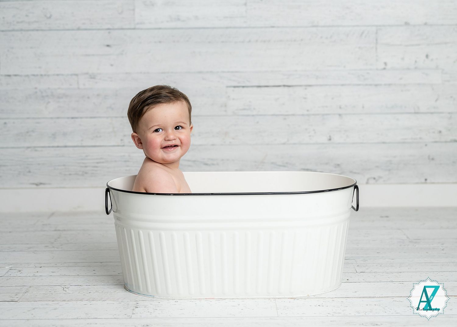 A happy baby sits in a white vintage-style wash basin against a light gray wooden backdrop.
