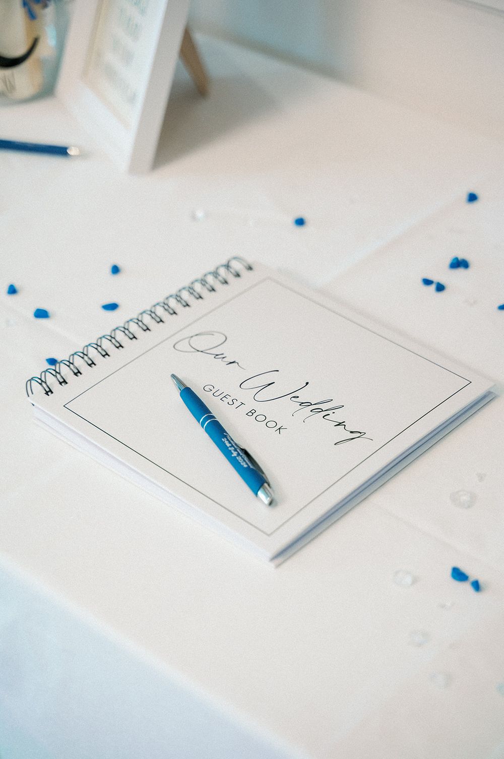 A guest book with a blue pen lies open on a white table with scattered blue confetti.
