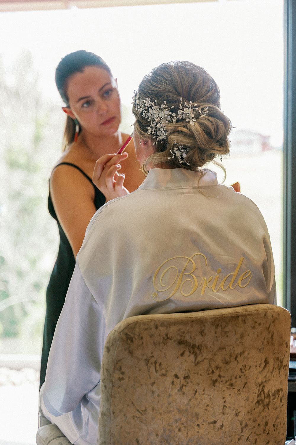 A person wearing a white robe with 'Bride' embroidery sits at a makeup station.