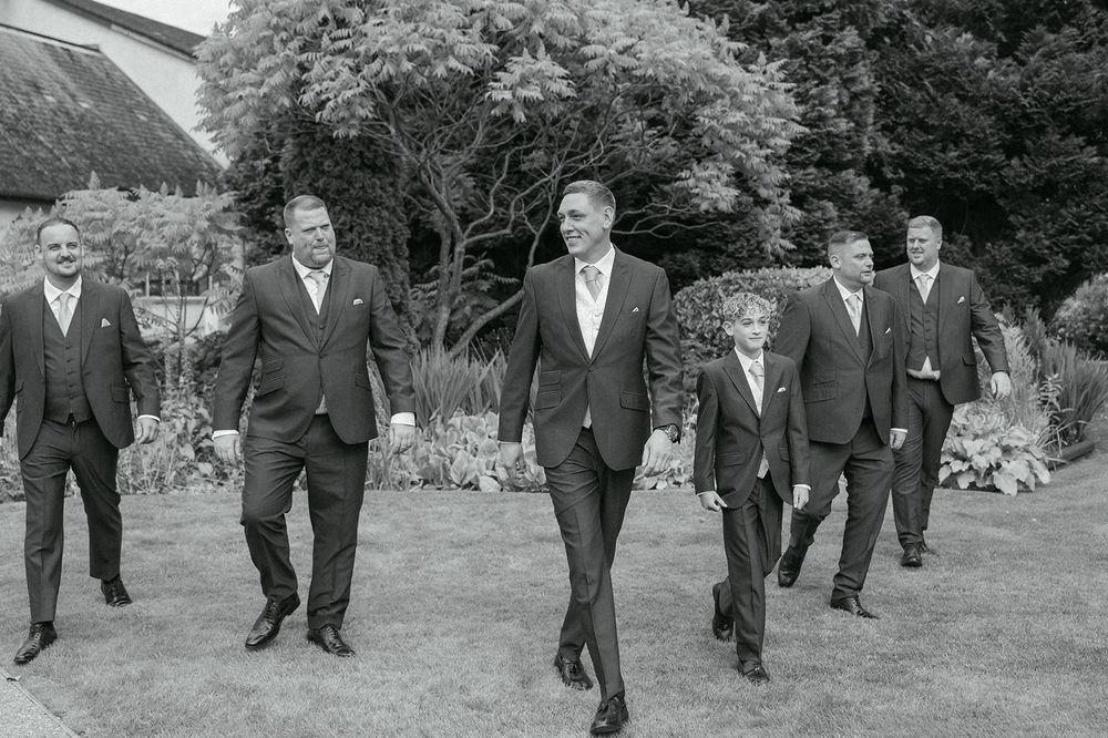 Groomsmen in formal attire walking in formation across a grassy lawn.