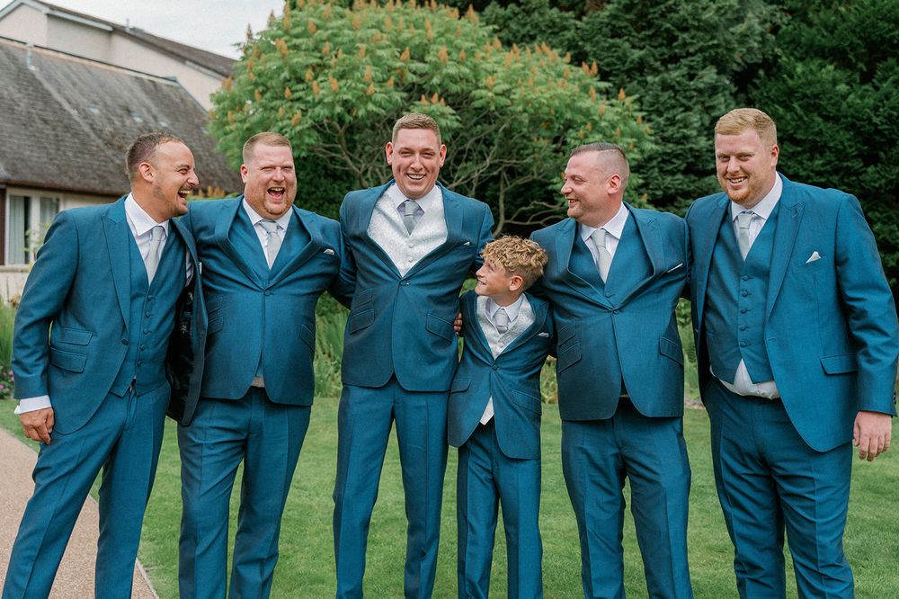 Groomsmen in matching blue suits pose together on a green lawn.