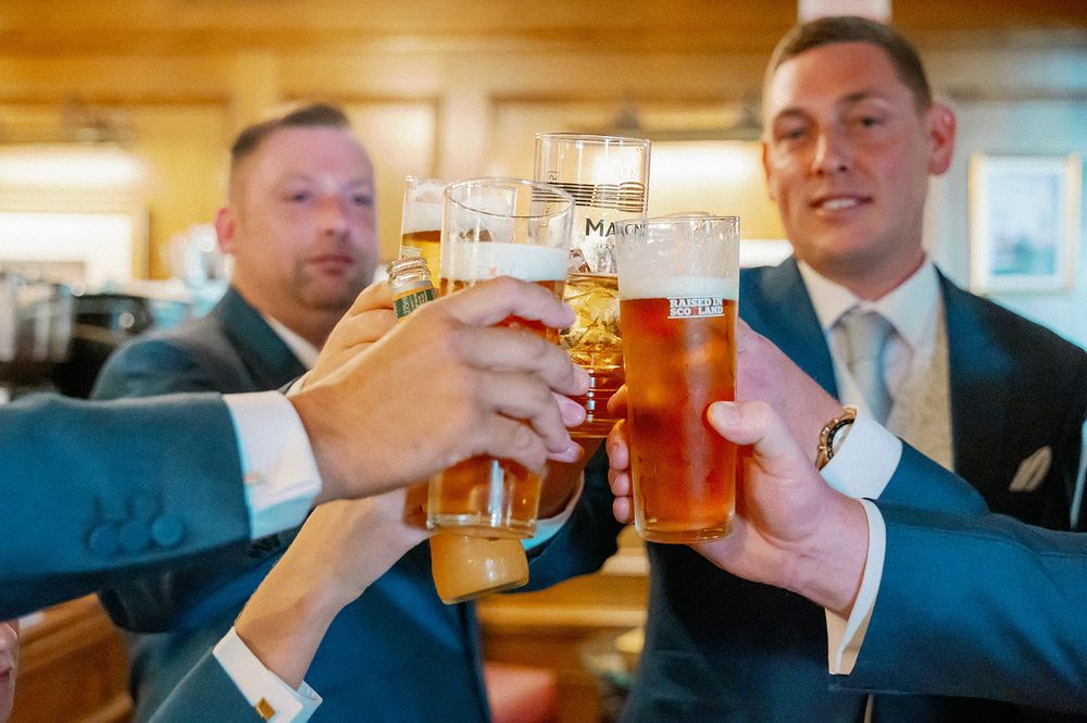 Wedding party members toasting with beer glasses in a warm-lit setting.