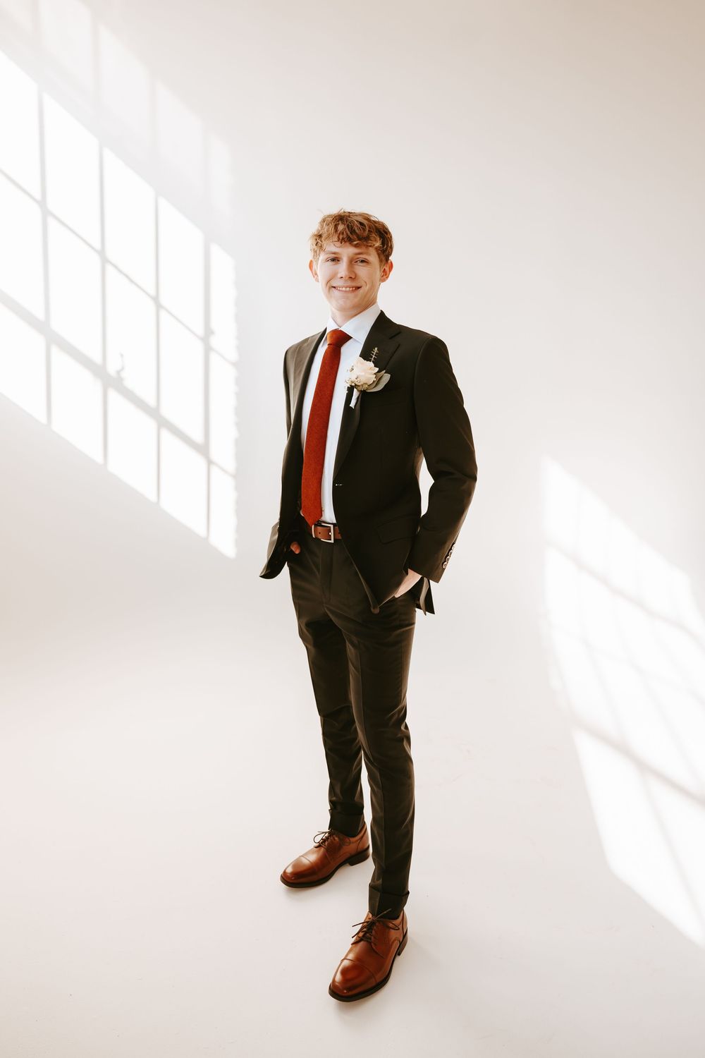Groom wearing dark suit with red tie and boutonniere poses against white wall with dramatic window lighting.