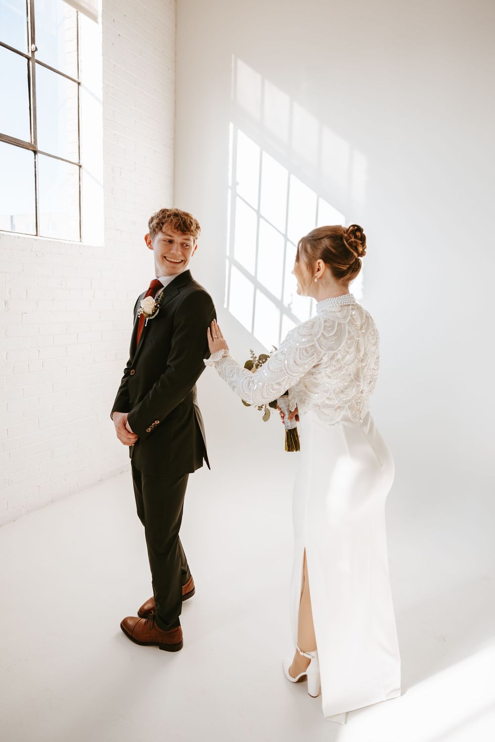 A couple in wedding attire stands in a minimalist white room with dramatic window lighting casting shadows.