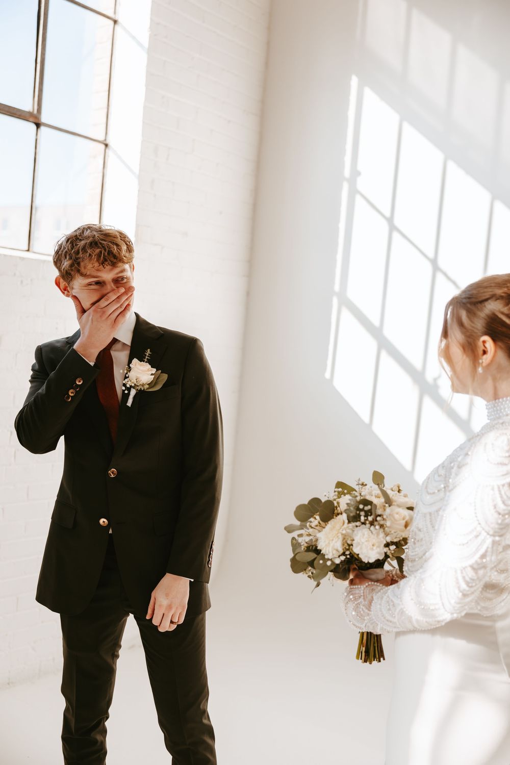 A candid wedding moment in a bright white room as an emotional groom reacts to seeing the bride holding her bouquet.