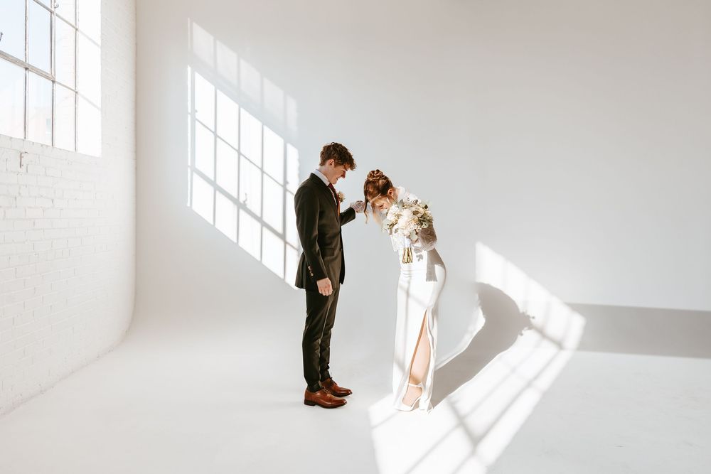 Two figures stand together in a bright white room with large windows creating dramatic natural lighting.