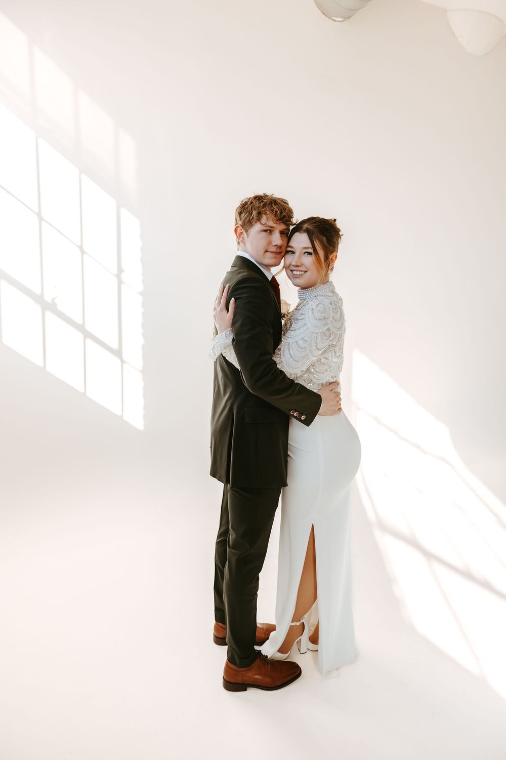 A couple embraces in a bright white room wearing wedding attire, with sunlight streaming through windows.