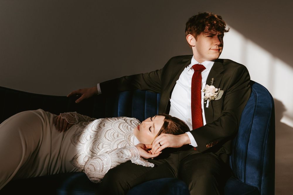 A couple in wedding attire rests together on a dark blue velvet couch in moody lighting.