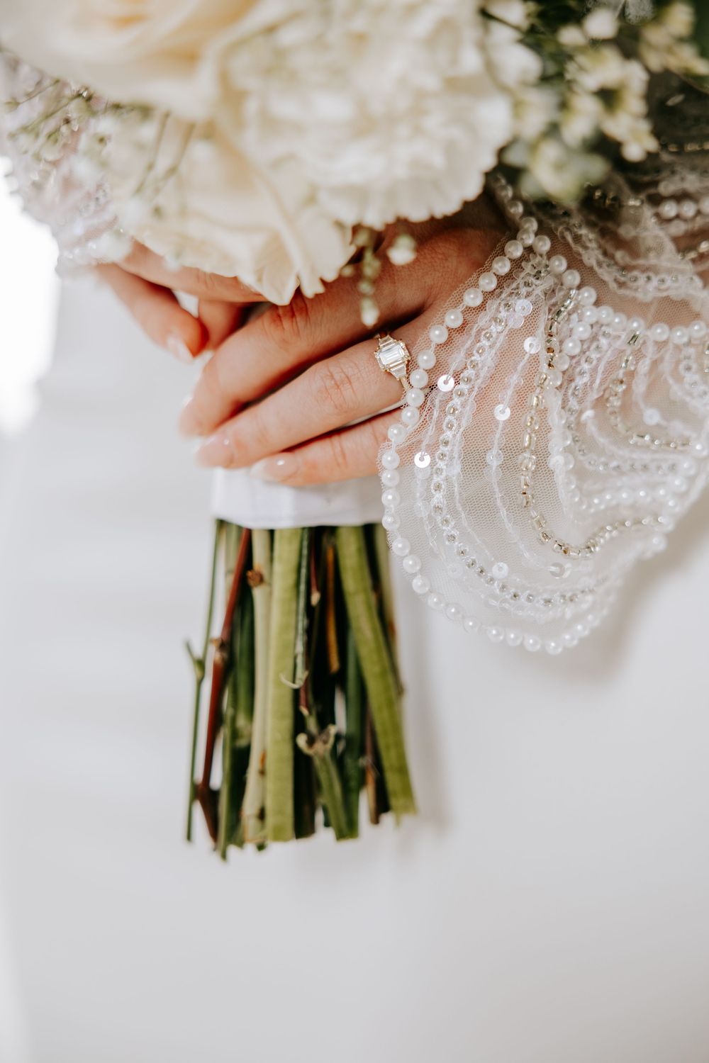 A close-up wedding photo showing hands holding a white floral bouquet with beaded dress detail in the frame.