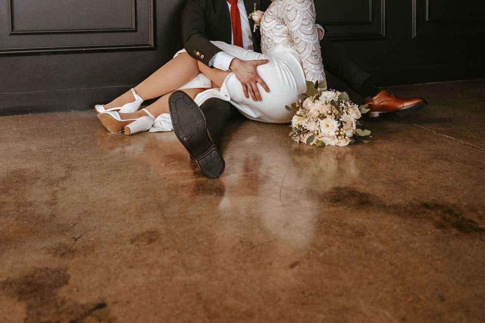 A white bridal bouquet lies on concrete floor next to feet wearing elegant bridal shoes and black dress shoes.
