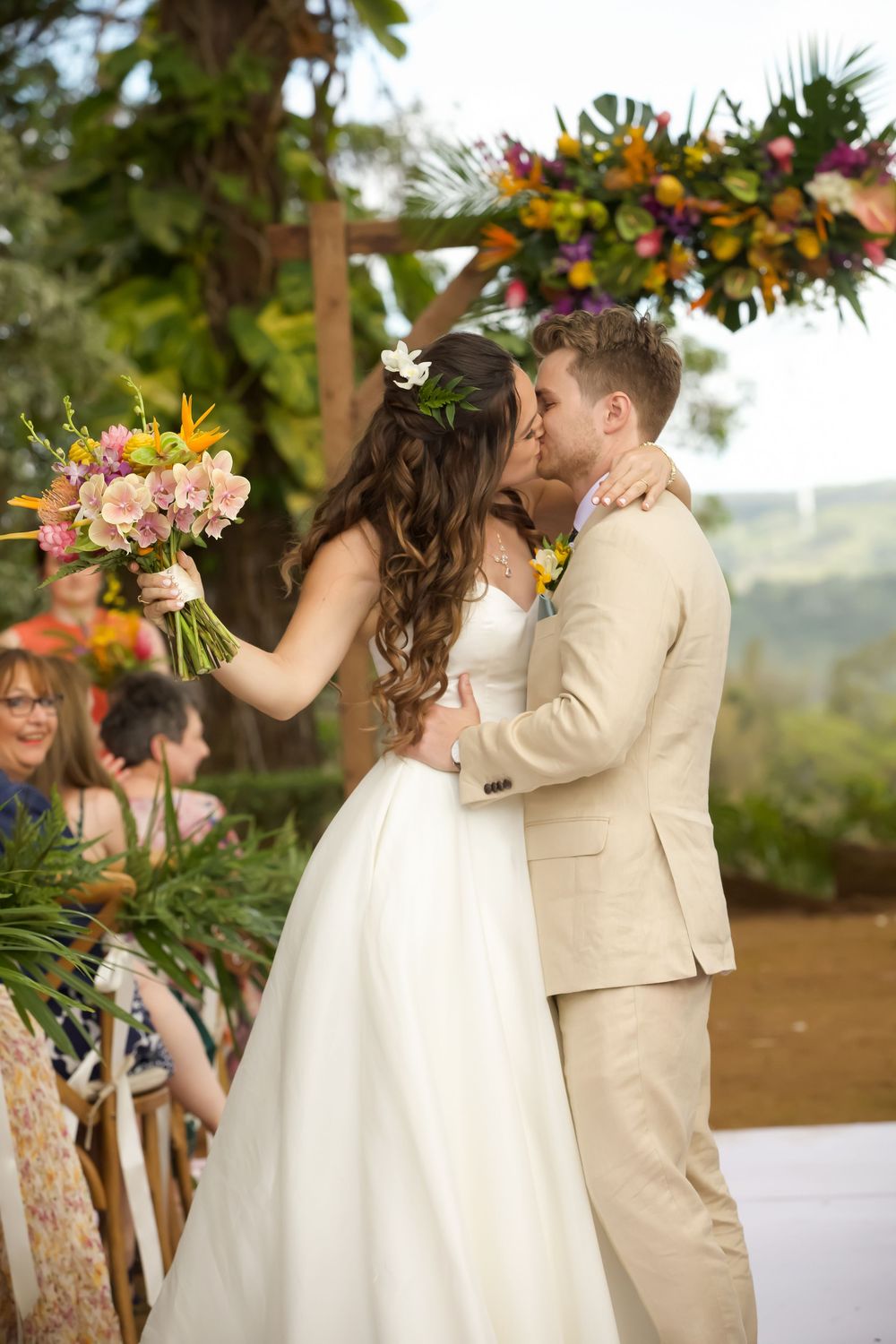 A romantic wedding kiss shared under a floral arch decorated with orange and pink blooms at an outdoor ceremony.