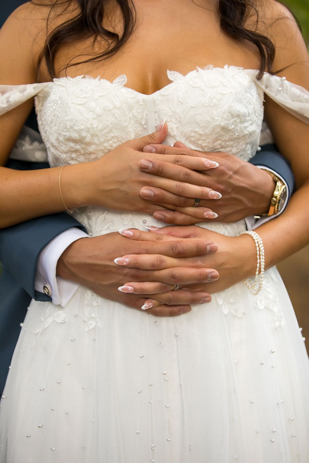 Hands with manicured nails embrace around a white lace wedding dress bodice adorned with delicate crystal details.
