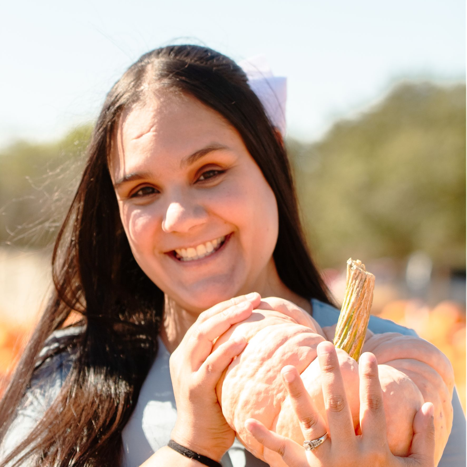 Someone with long dark hair holds a light pink pumpkin outdoors on a sunny autumn day.