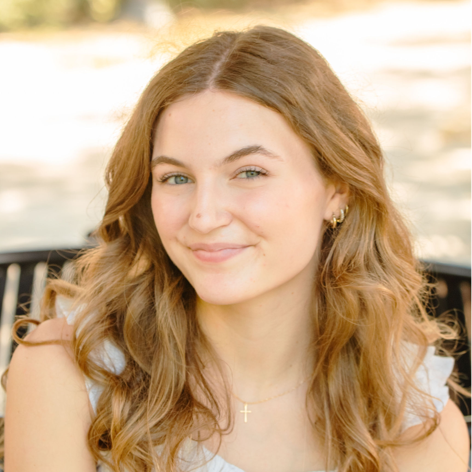 A person with long wavy honey blonde hair smiles warmly while sitting outdoors in natural lighting.