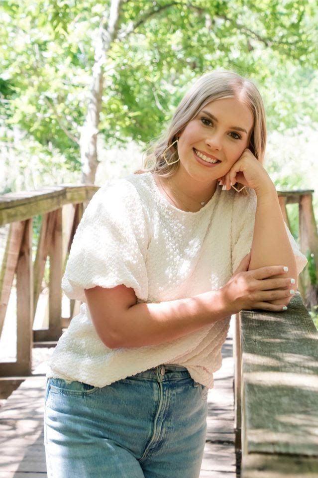 A person in a white lace top and blue jeans poses casually outdoors in a sunlit wooded setting.
