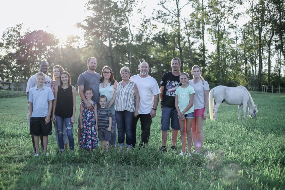 A group of people stand together in a grassy field at sunset with a white horse grazing in the background.