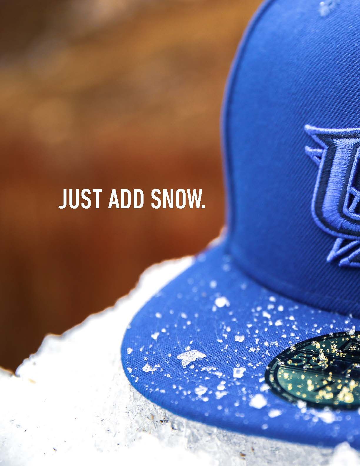 Blue fitted baseball cap with logo resting in snow with text Just Add Snow on blurred background.