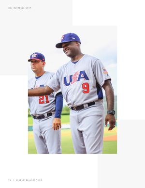 Two USA Baseball coaches in gray uniforms numbered 21 and 9 standing on baseball field.