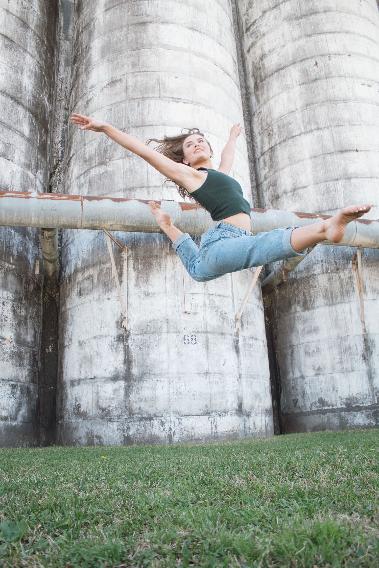 Dynamic dancer performing a graceful sideways leap sequence in front of concrete silos on grassy lawn.