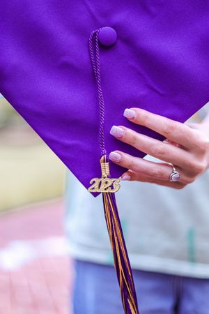 Close-up of a purple graduation cap tassel with gold 2023 charm and manicured hand with engagement ring.