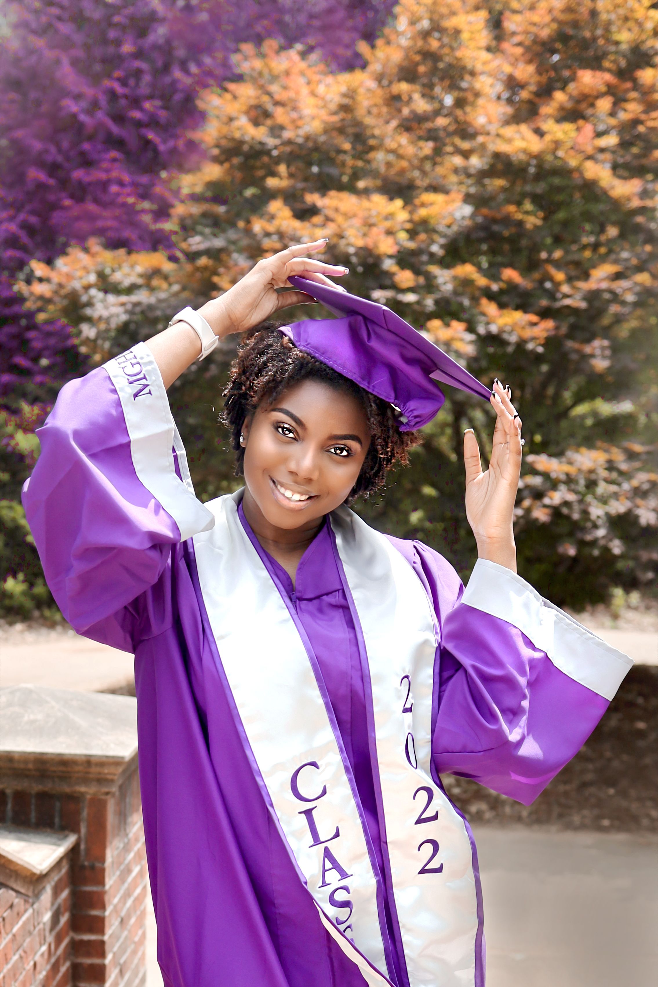 Graduate in purple and white academic regalia poses joyfully against colorful autumn foliage.