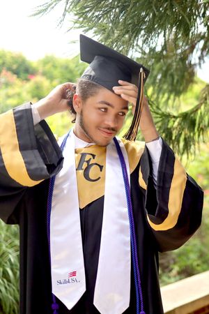 A graduate adjusts their cap while wearing traditional commencement regalia with blue and gold colors outdoors.
