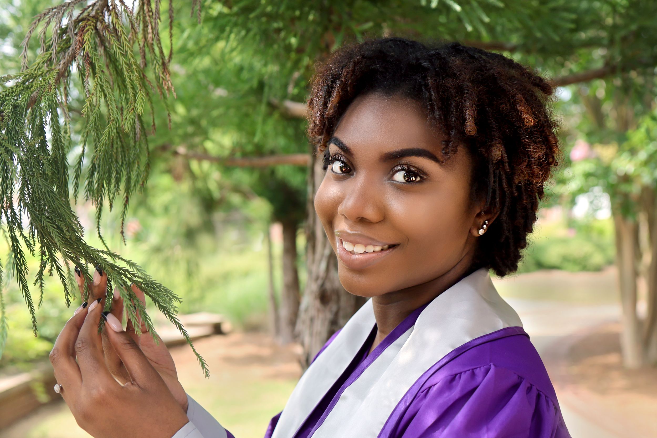 Graduate in purple academic regalia stands beneath pine trees on a sunny day in an outdoor setting.