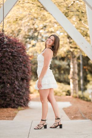 A woman in a white summer dress poses outdoors against a soft bokeh background with autumn foliage.