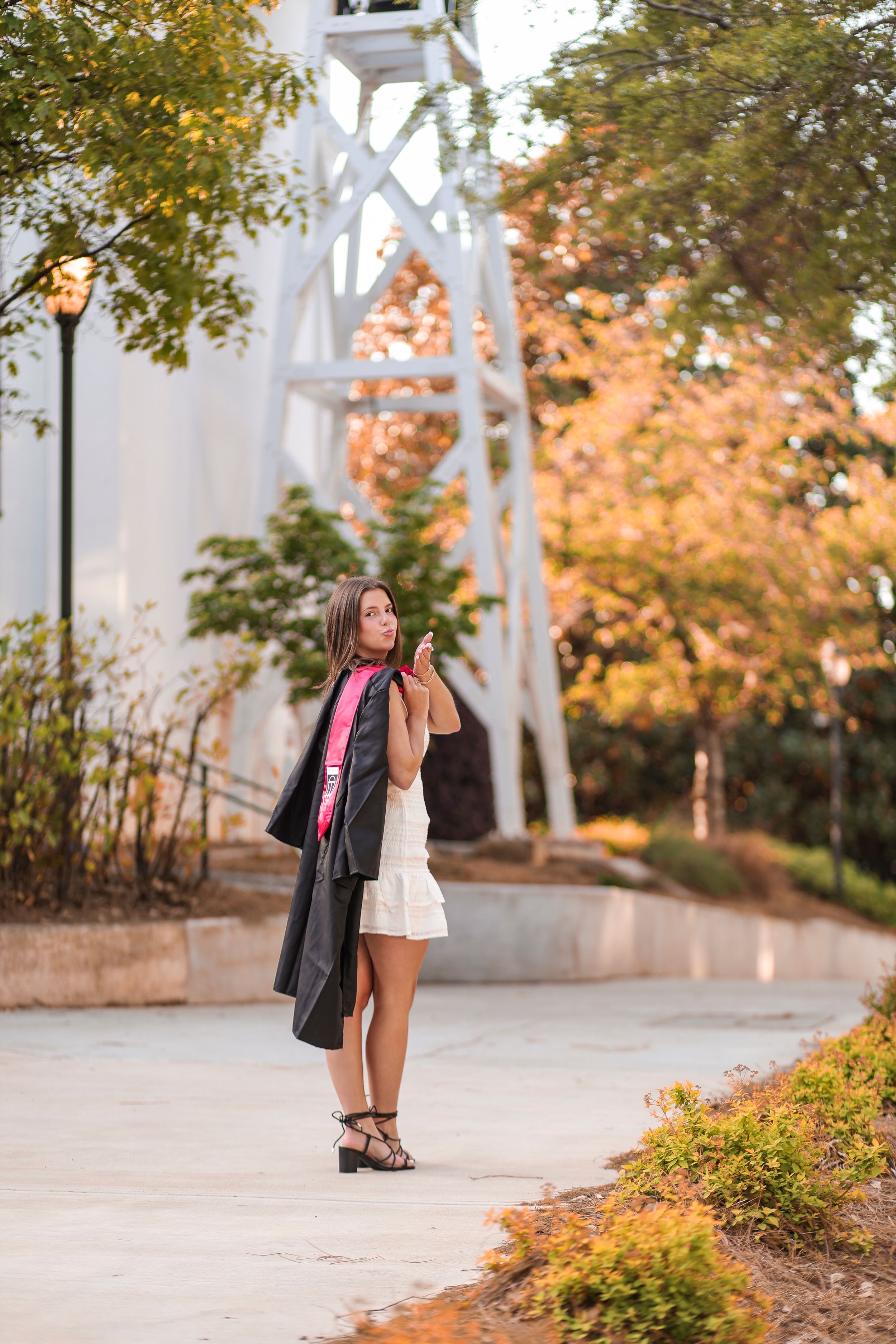 Figure in a white dress stands on a path surrounded by autumn foliage near a white architectural structure.