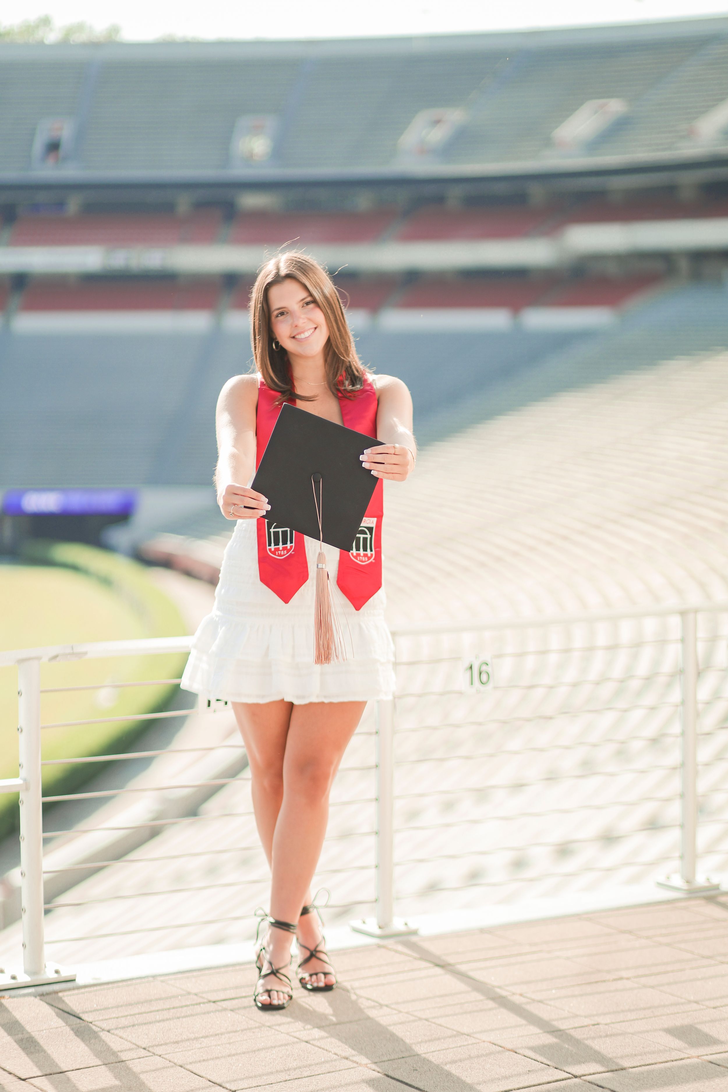 A person in a red and white outfit stands holding a graduation cap on outdoor stadium stairs in sunlight.