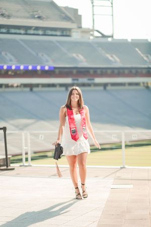 Student in white dress and red sash walks on steps of empty stadium in bright sunlight.