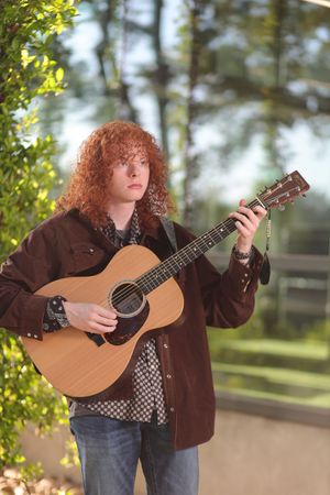 Musician with curly red hair plays acoustic guitar outdoors while wearing a brown jacket and jeans.