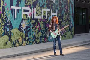 A musician with colorful hair plays guitar in front of a vibrant street art mural with 'TRUTH' text on the wall.