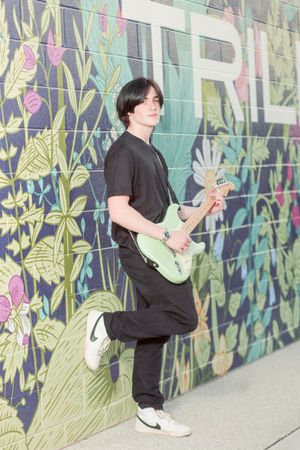 Musician in black attire playing mint green guitar against colorful floral mural wall.