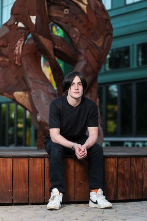 Someone in all black clothing sits on a wooden bench in front of a large metal sculpture on a city street.