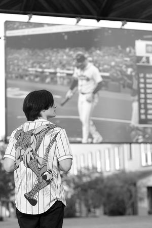 Young fan in pinstripe baseball jersey watches a game on stadium screen while holding a drink in black and white.
