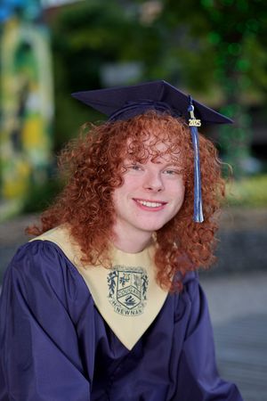Graduate wearing navy cap and gown with curly red hair poses outdoors against blurred background at commencement.